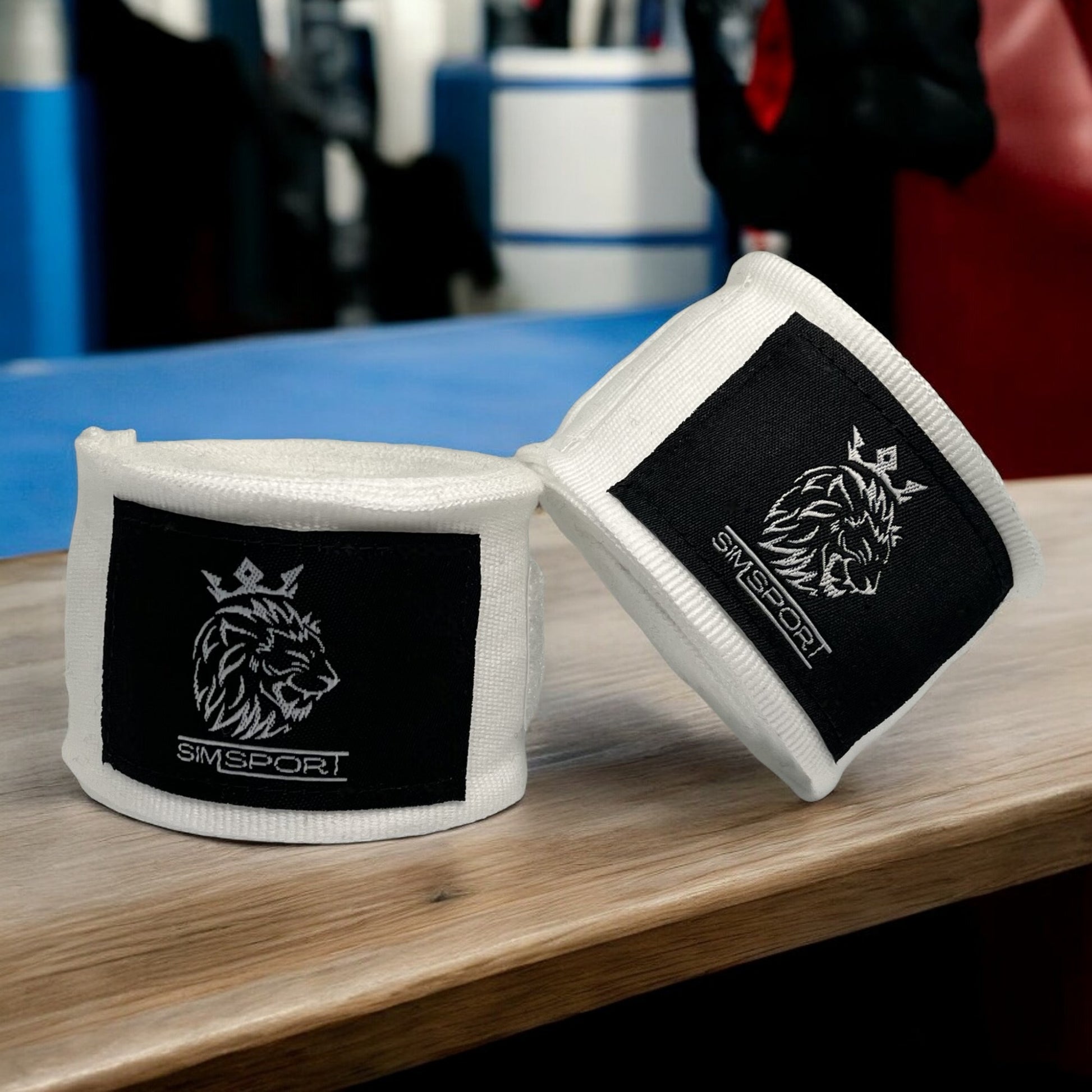 Bandes de boxe SIMSPORT couleur blanc posées sur une table en bois avec un ring de boxe en arrière plan