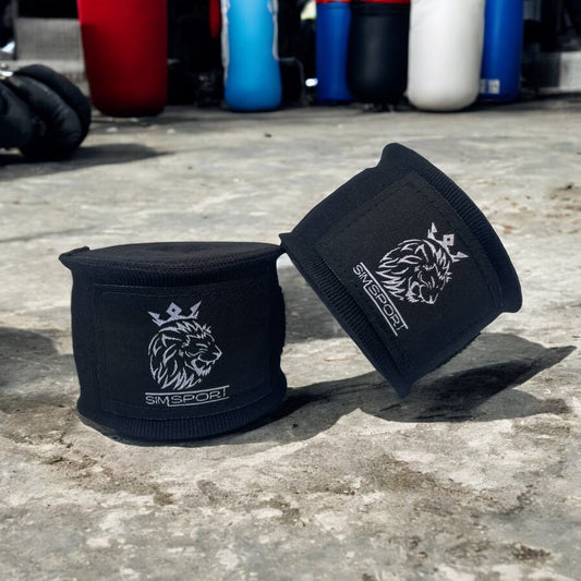 Bandes de boxe SIMSPORT couleur noir posées sur un sol en béton salle de boxe en arrière plan, sacs de frappe