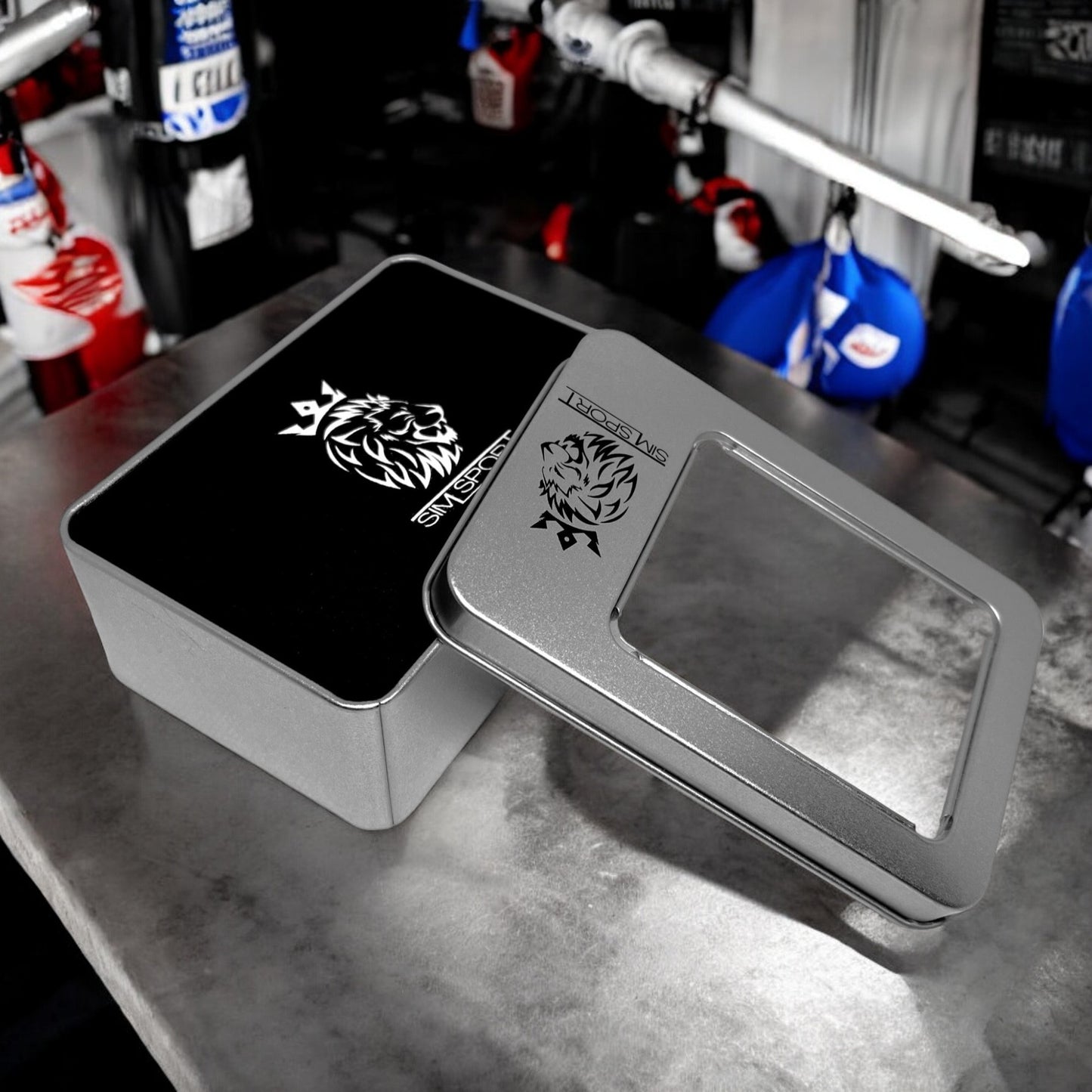 Pad de protection mains (métacarpes) posés sur une table grise, offert avec sa boîte métallique, couleur noir, sacs de frappe boxe en arrière plan