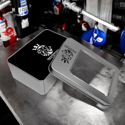 Pad de protection mains (métacarpes) posés sur une table grise, offert avec sa boîte métallique, couleur noir, sacs de frappe boxe en arrière plan